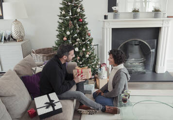 Husband giving Christmas gift to wife by tree in living room - CAIF30281