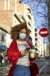 Young woman wearing face mask using mobile phone while standing with coffee cup in city - VEGF03693