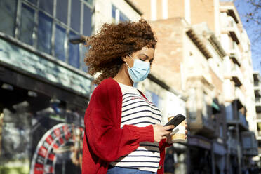 Curly hair woman wearing sanitary mask holding coffee cup while using mobile phone standing in city - VEGF03694