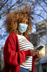 Young curly hair woman wearing protective face mask using mobile phone while standing outdoors - VEGF03695