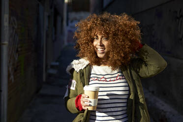 Lächelnde Frau mit Hand im Haar, die eine Kaffeetasse hält, während sie auf einem Fußweg steht - VEGF03697