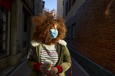 Brown curly hair woman wearing protective face mask looking away while standing with coffee cup on footpath - VEGF03698