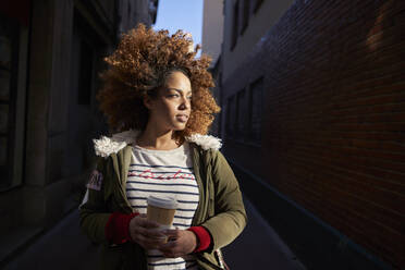 Young woman with disposable coffee cup looking away while standing on footpath - VEGF03699