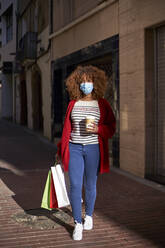 Junge Frau mit Gesichtsschutz, die eine Kaffeetasse und eine Einkaufstasche in der Hand hält, während sie auf einem Fußweg geht - VEGF03708