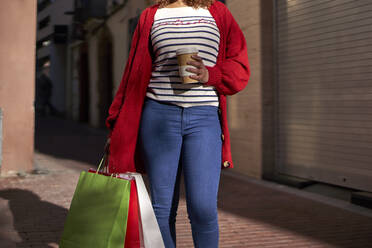Frau hält Einweg-Kaffeebecher und Einkaufstasche, während sie auf dem Fußweg steht - VEGF03710