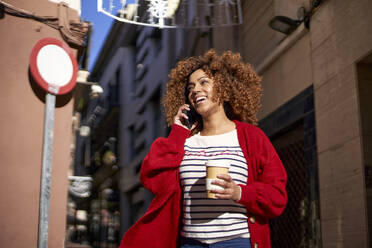 Lächelnde junge Frau mit Einweg-Kaffeebecher, die mit ihrem Handy telefoniert, während sie vor einem Gebäude steht - VEGF03715