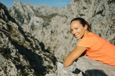 Mittlere erwachsene Frau lächelt, während sie sich auf einem Felsen am Cares Trail im Nationalpark Picos De Europe, Asturien, Spanien, ausruht - DMGF00459