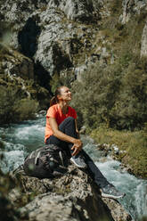 Mittlere erwachsene Frau, die wegschaut, während sie am Fluss am Cares Trail im Nationalpark Picos De Europe, Asturien, Spanien, sitzt - DMGF00474