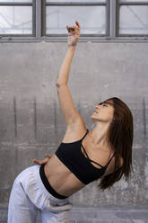 Tänzerin mit erhobenem Arm tanzt gegen eine graue Wand in einer verlassenen Fabrik - VEGF03730
