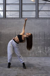 Mid adult female dancer stretching while standing against gray wall in abandoned factory - VEGF03731