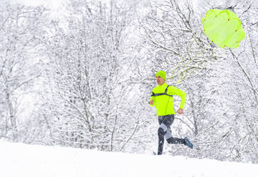 Männlicher Sportler mit Fallschirm, der im Winter auf Schnee im Wald läuft - STSF02817