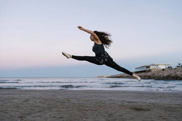 Anmutige Balletttänzerin mit gespreizten Beinen beim Üben am Strand bei Sonnenuntergang - EGAF01653