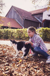 Girl playing with cat while crouching on dry leaf path - DHEF00517