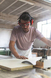 Young male carpenter cutting wood using table saw while working in workshop - JAQF00217