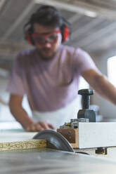 Male carpenter cutting wood with table saw while working in workshop - JAQF00218