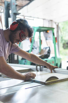 Junger männlicher Handwerker schneidet Holz mit einer Tischsäge in einer Werkstatt - JAQF00219