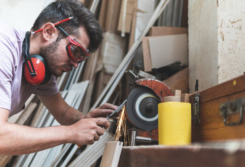 Konzentrierter männlicher Tischler beim Schärfen von Werkzeugen auf einer Schleifmaschine in einer Werkstatt - JAQF00229