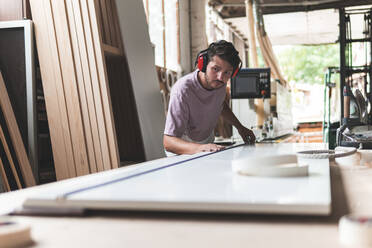 Male carpenter measuring plank using tape measure on workbench in workshop - JAQF00231