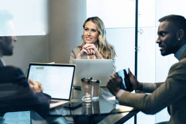 Smiling businesswoman with hand on chin listening to businessman while sitting in meeting at office - JSRF01357