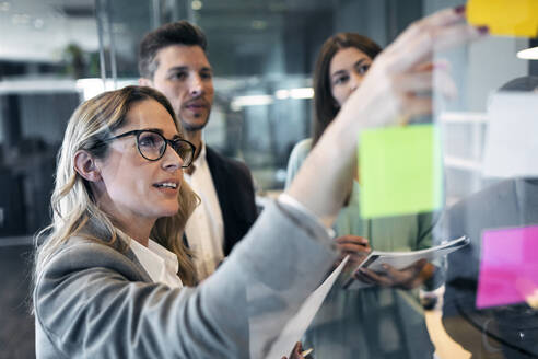 Businesswoman reading adhesive note on glass wall while standing with colleague at office - JSRF01368