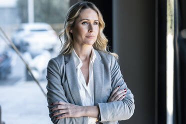 Thoughtful businesswoman looking away while standing with arms crossed in office - JSRF01372