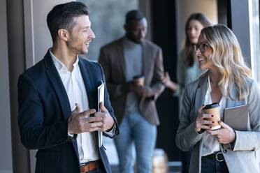 Smiling business people discussing while standing with colleague in background at office - JSRF01377