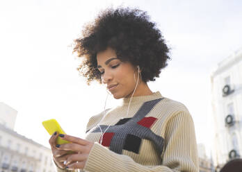 Young woman wearing in-ear headphones using mobile phone while standing outdoors - JCCMF01071