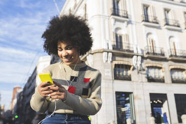 Smiling afro woman using smart phone while standing in city - JCCMF01073