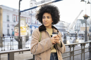 Afro woman with disposable cup looking away while leaning on railing - JCCMF01107