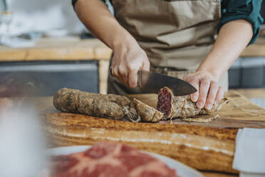 Female chef cutting salami on cutting board while standing in kitchen - MFF06955