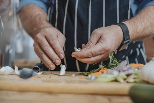 Male chef wearing apron peeling garlic while doing preparation standing in kitchen - MFF06961