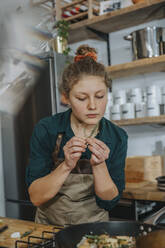 Young chef smelling thyme leaf while standing in kitchen - MFF06990