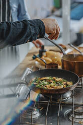 Chef sprinkling vegetable leaf over food in frying pan while standing by colleague in kitchen - MFF06996