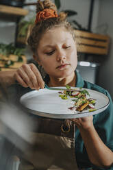 Young chef arranging vegetable with tongs in plate while standing in kitchen - MFF07024