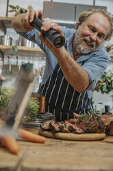 Smiling chef seasoning pepper on grilled tomahawk steak while standing in kitchen - MFF07028