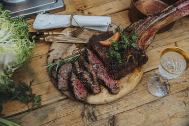 Tomahawk steak garnished with vegetables on wooden plate by drink in kitchen - MFF07029