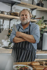 Confident mature chef holding kitchen knife while standing with arms crossed in kitchen - MFF07037