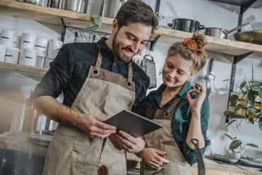 Female chef gesturing OK sign while listening to video call on digital tablet by colleague in kitchen - MFF07041