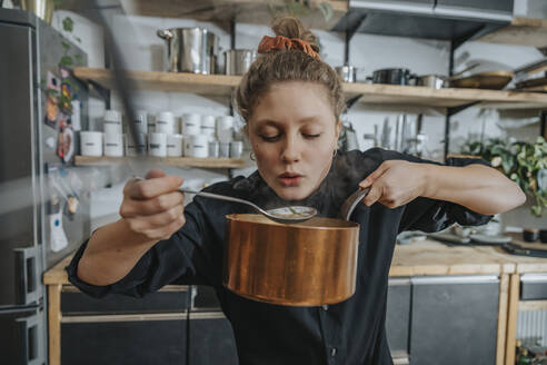 Young female chef tasting Broth soup while standing in kitchen - MFF07055