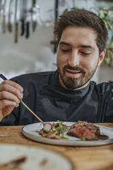 Smiling chef arranging cooked meat and vegetables in plate while standing in kitchen - MFF07064