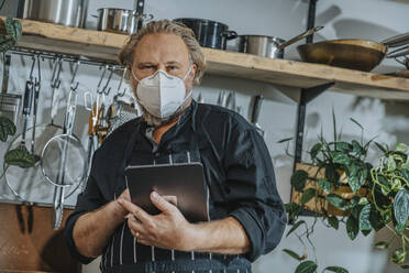 Mature expertise wearing protective face mask using digital tablet while standing at kitchen - MFF07073