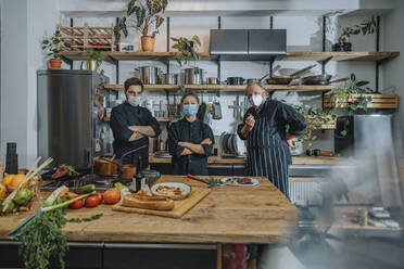 Confident chef wearing protective face mask staring while standing at kitchen - MFF07076