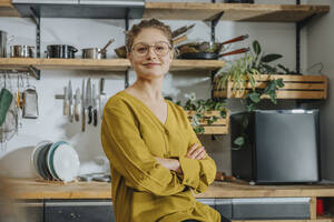 Confident young woman smiling while sitting with arms crossed in kitchen - MFF07087