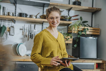 Junge Frau mit Brille, die lächelnd ein digitales Tablet in der Küche benutzt - MFF07088