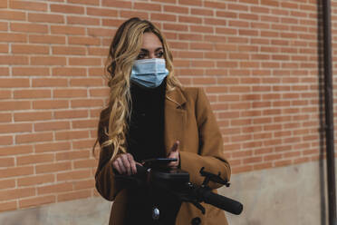 Young woman in protective face mask standing with electric push scooter against brick wall - FMOF01318