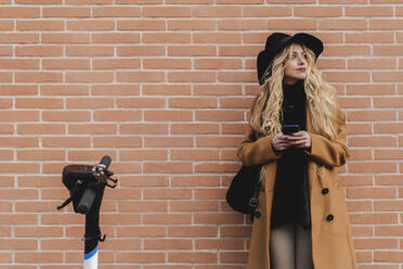 Frau im Mantel, die ein Smartphone benutzt, während sie an einer Mauer steht - FMOF01322