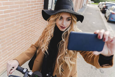 Frau mit blondem Haar, die ein Selfie auf einem Fußweg an einer Mauer macht - FMOF01324