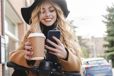 Happy young woman using smart phone while holding reusable coffee cup - FMOF01326