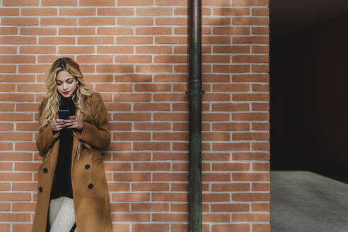 Junge Frau, die ihr Smartphone benutzt, während sie Musik gegen eine Backsteinmauer hört - FMOF01334