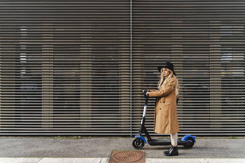 Woman with electric push scooter standing against metallic wall - FMOF01342